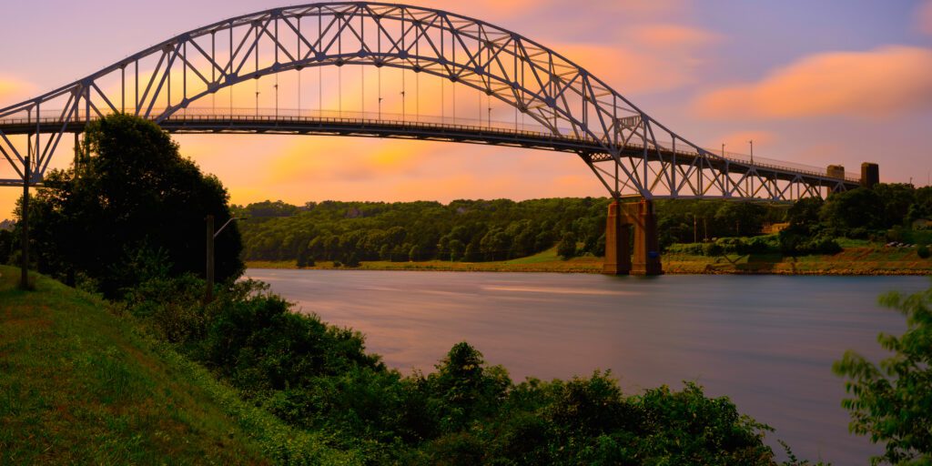 Sagamore Bridge at Sunrise over Cape Cod Canal with floating orange colored clouds and green bushes and tree on the riverbank on Cape Cod, Massachusetts, USA, Long Exposure photo. Exclusive Yachts